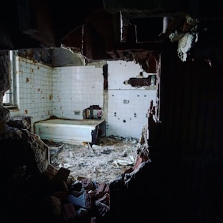 A dilapidated bathroom with broken walls and debris scattered on the floor. The room has white tiles, some of which are damaged, and an old bathtub. The lighting is dim, casting shadows and creating a somber atmosphere.