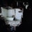 A dilapidated bathroom with broken walls and debris scattered on the floor. The room has white tiles, some of which are damaged, and an old bathtub. The lighting is dim, casting shadows and creating a somber atmosphere.