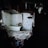 A dilapidated bathroom with broken walls and debris scattered on the floor. The room has white tiles, some of which are damaged, and an old bathtub. The lighting is dim, casting shadows and creating a somber atmosphere.