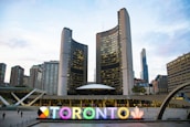 A modern cityscape featuring a large, brightly lit sign spelling 'TORONTO' with a multi-colored letter and a maple leaf symbol. Behind the sign, tall office buildings, including a distinctive curved twin tower structure, are visible under a partly cloudy sky.
