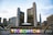 A modern cityscape featuring a large, brightly lit sign spelling 'TORONTO' with a multi-colored letter and a maple leaf symbol. Behind the sign, tall office buildings, including a distinctive curved twin tower structure, are visible under a partly cloudy sky.