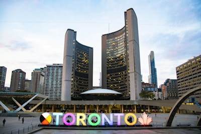 A modern cityscape featuring a large, brightly lit sign spelling 'TORONTO' with a multi-colored letter and a maple leaf symbol. Behind the sign, tall office buildings, including a distinctive curved twin tower structure, are visible under a partly cloudy sky.