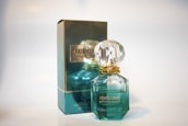 A perfume bottle and its packaging are displayed. The bottle has a gem-like, faceted stopper and is filled with a light green liquid. The box is similarly colored in a reflective teal with gold accents and features the branding.