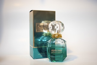 A perfume bottle and its packaging are displayed. The bottle has a gem-like, faceted stopper and is filled with a light green liquid. The box is similarly colored in a reflective teal with gold accents and features the branding.
