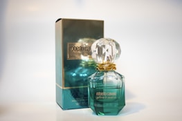 A perfume bottle and its packaging are displayed. The bottle has a gem-like, faceted stopper and is filled with a light green liquid. The box is similarly colored in a reflective teal with gold accents and features the branding.