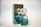A perfume bottle and its packaging are displayed. The bottle has a gem-like, faceted stopper and is filled with a light green liquid. The box is similarly colored in a reflective teal with gold accents and features the branding.