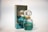 A perfume bottle and its packaging are displayed. The bottle has a gem-like, faceted stopper and is filled with a light green liquid. The box is similarly colored in a reflective teal with gold accents and features the branding.