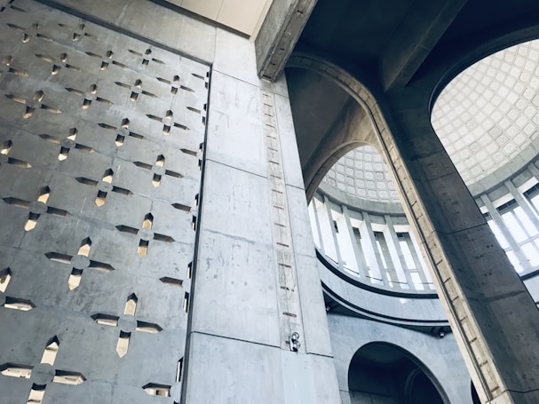 Interior view of a concrete building showcasing a blend of brutalist and modern architectural elements. The structure features high ceilings with large arches, numerous angular cutouts in the walls, and a well-lit circular dome. The design incorporates geometric patterns and extensive use of raw concrete.