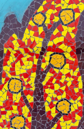 A colorful mosaic composed of irregularly shaped tiles predominantly in shades of red, yellow, and dark maroon. The tiles are arranged in a pattern that includes circular and linear designs, which could resemble abstract forms or symbols. The background features a light blue section in the upper corners, providing contrast to the vivid primary colors.