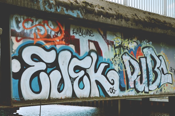 Graffiti art with large, bold letters covers a concrete wall. The foreground features prominent, stylized letters in white outlined with black and blue. The background includes layers of overlapping tags and images in bright colors like red and yellow, showcasing a variety of artistic styles.