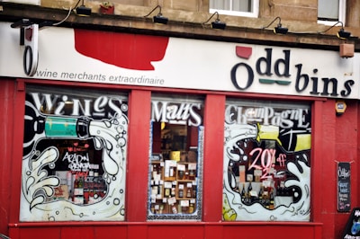 A storefront with large glass windows displaying a variety of wine bottles. The exterior features vibrant artwork of wine bottles pouring liquid, with bold text advertising wines, malts, and champagne. A sign above the windows reads 'Oddbins,' and there is a chalkboard to the side with some writing.