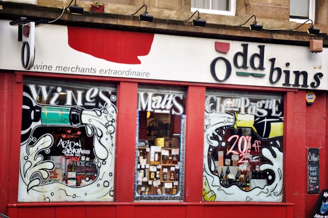 A storefront with large glass windows displaying a variety of wine bottles. The exterior features vibrant artwork of wine bottles pouring liquid, with bold text advertising wines, malts, and champagne. A sign above the windows reads 'Oddbins,' and there is a chalkboard to the side with some writing.