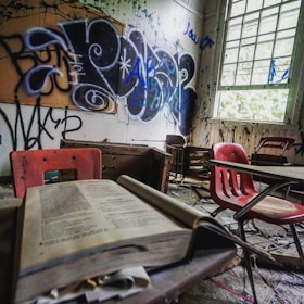 A dilapidated classroom with overturned desks and chairs scattered around. Graffiti covers the old, peeling walls, and an open textbook rests on a desk in the foreground. Light filters through a large window, casting shadows across the room. The air seems heavy with neglect and abandonment.