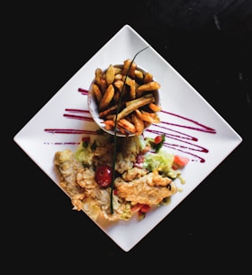 A square white plate displays a serving of crispy golden-brown fries in a small cup, accompanied by fried chicken pieces on a bed of lettuce and diced colorful vegetables. The presentation includes artistic strokes of purple sauce across the plate.