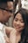 A close-up of two people smiling at each other, with natural lighting coming through the trees in the blurred background. One person has short hair and glasses, while the other has long hair and is looking downward with a warm smile. They appear to be in a peaceful, wooded area.