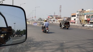 A busy urban street with various vehicles including an auto rickshaw and a motorcycle carrying two people. Buildings with commercial signage line the road, and a side-view mirror of a nearby vehicle reflects part of the scene.