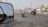 A busy urban street with various vehicles including an auto rickshaw and a motorcycle carrying two people. Buildings with commercial signage line the road, and a side-view mirror of a nearby vehicle reflects part of the scene.