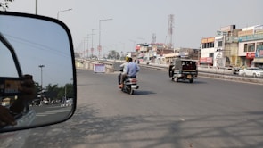 A busy urban street with various vehicles including an auto rickshaw and a motorcycle carrying two people. Buildings with commercial signage line the road, and a side-view mirror of a nearby vehicle reflects part of the scene.