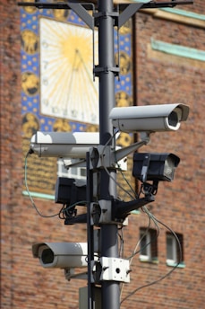 Several surveillance cameras are mounted on a metal pole, with a detailed background featuring a decorative brick wall and a large clock or sundial with a sun motif.