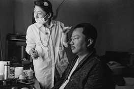 A black and white image featuring two individuals indoors, where one is applying makeup to the other. The person applying makeup wears a mask and holds a brush, appearing focused on their task. Various makeup supplies or cosmetic items are visible on a nearby table.