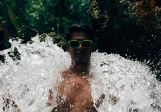 A person wearing green sunglasses stands amid a waterfall, with water splashing around. The background features lush green foliage and a dark, rocky surface. The scene conveys a sense of adventure and interaction with nature.
