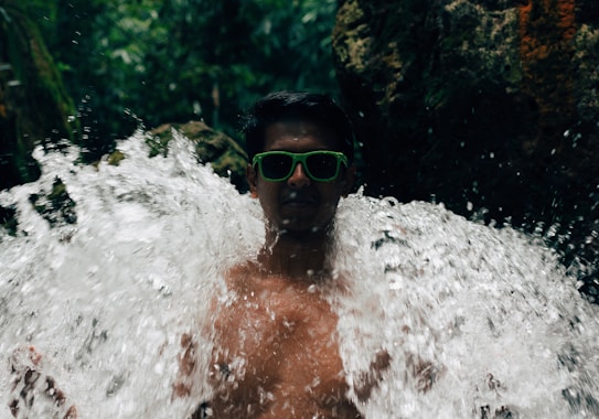 A person wearing green sunglasses stands amid a waterfall, with water splashing around. The background features lush green foliage and a dark, rocky surface. The scene conveys a sense of adventure and interaction with nature.