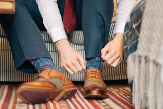 A person is sitting on a striped couch while adjusting colorful socks with a playful pattern. They are wearing a white shirt and gray trousers. The person's hands are focused on securing the socks within a pair of brown leather brogue shoes. The floor is covered with a striped rug.