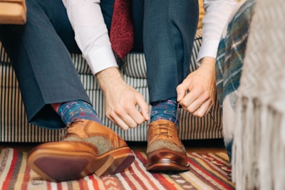 A person is sitting on a striped couch while adjusting colorful socks with a playful pattern. They are wearing a white shirt and gray trousers. The person's hands are focused on securing the socks within a pair of brown leather brogue shoes. The floor is covered with a striped rug.