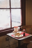 A wooden table near a window with red frames holds a plate of food including fried items and coleslaw. There is a small bottle of Coca-Cola, disposable utensils, a menu, and sauce packets on the table. The scene appears to be inside a casual dining establishment with natural light coming through the window.