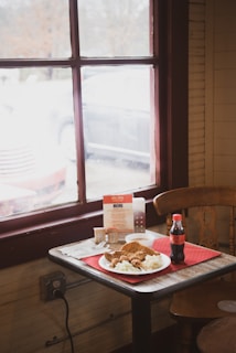 A wooden table near a window with red frames holds a plate of food including fried items and coleslaw. There is a small bottle of Coca-Cola, disposable utensils, a menu, and sauce packets on the table. The scene appears to be inside a casual dining establishment with natural light coming through the window.