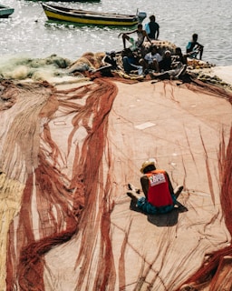 Several individuals are working with fishing nets spread out along a waterfront. The nets are red and tan, and a person in a red shirt and hat is seated in the foreground, focusing on the task. The scene is set against a backdrop of water where a few boats are visible.