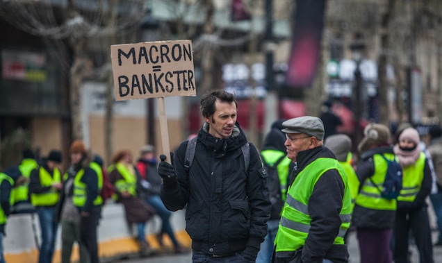 A group of people is engaged in a protest. One individual in the foreground holds a sign with a political message. Several people are wearing high-visibility yellow vests, indicating participation in a demonstration or protest.