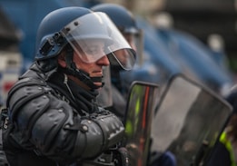 A person is wearing riot gear, including a blue helmet with a clear face shield and black padded armor. They hold a transparent riot shield with reflections visible on its surface. The background is slightly blurred, showing more individuals in similar attire.