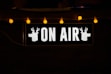 A black rectangular sign with the words 'ON AIR' in bold, white letters. On either side of the text, there are silhouettes of deer heads. Above the sign, several warm yellow fairy lights provide a soft, ambient glow.