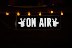 A black rectangular sign with the words 'ON AIR' in bold, white letters. On either side of the text, there are silhouettes of deer heads. Above the sign, several warm yellow fairy lights provide a soft, ambient glow.