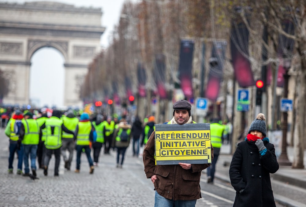 standing man wearing yellow and black signage near people during daytime