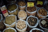 Various types of dried herbs and roots are displayed in large, open plastic bags, each containing a different kind of herb or root. Some bags have labels with prices written in Chinese characters. The image is likely taken in an herbal market or shop.