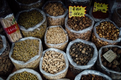 Various types of dried herbs and roots are displayed in large, open plastic bags, each containing a different kind of herb or root. Some bags have labels with prices written in Chinese characters. The image is likely taken in an herbal market or shop.