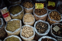Various types of dried herbs and roots are displayed in large, open plastic bags, each containing a different kind of herb or root. Some bags have labels with prices written in Chinese characters. The image is likely taken in an herbal market or shop.