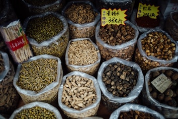 Various types of dried herbs and roots are displayed in large, open plastic bags, each containing a different kind of herb or root. Some bags have labels with prices written in Chinese characters. The image is likely taken in an herbal market or shop.