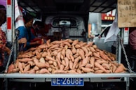 A large pile of sweet potatoes is displayed on the back of a truck, surrounded by people selecting or purchasing them. There is a handwritten sign in Chinese on the side, and the truck is parked in an urban environment with other vehicles visible in the background.