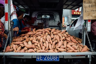 A large pile of sweet potatoes is displayed on the back of a truck, surrounded by people selecting or purchasing them. There is a handwritten sign in Chinese on the side, and the truck is parked in an urban environment with other vehicles visible in the background.
