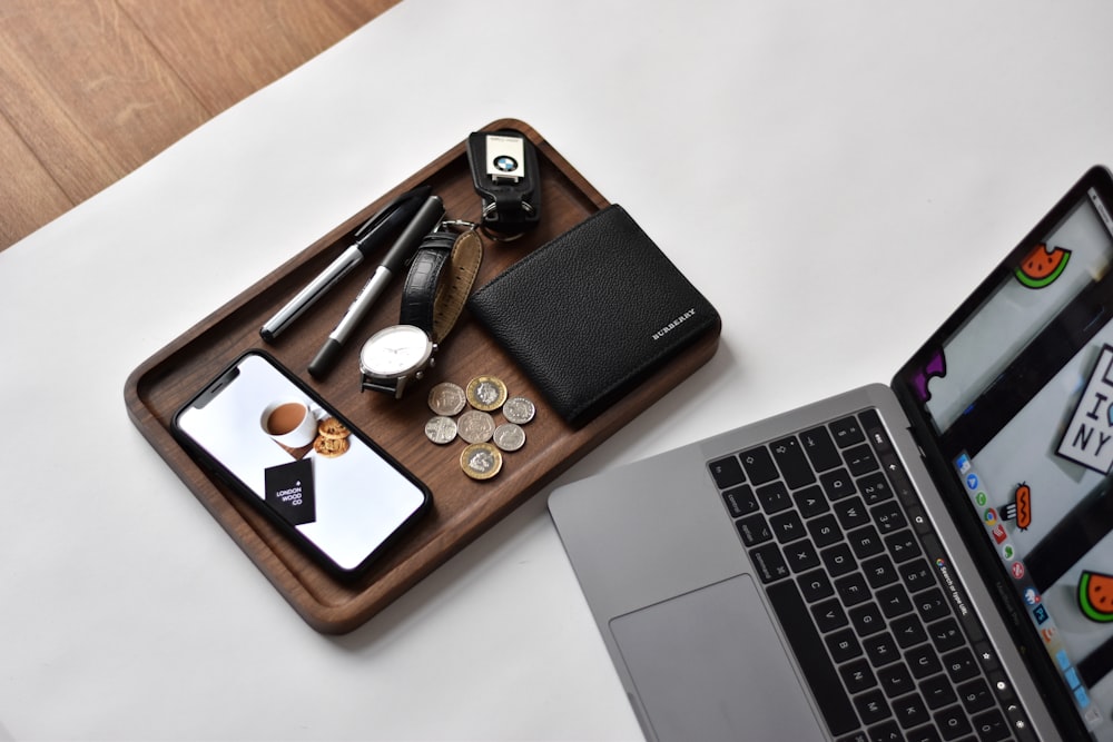 turned-on laptop beside phone, wallet, watch, pens, and coins on top of wooden tray