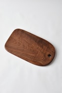 A wooden cutting board with a smooth finish and rounded edges lies on a plain white surface. The wood grain and rich brown color are prominent, with a circular hole near one end.