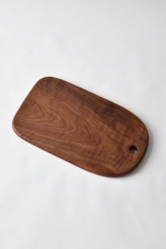 A wooden cutting board with a smooth finish and rounded edges lies on a plain white surface. The wood grain and rich brown color are prominent, with a circular hole near one end.