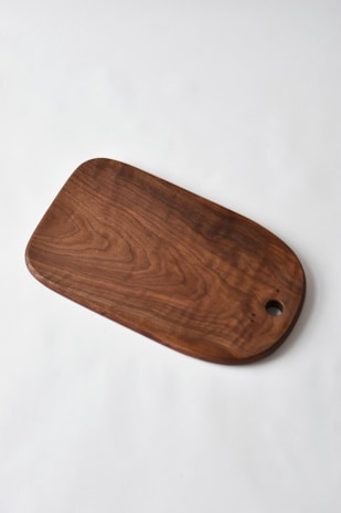 A wooden cutting board with a smooth finish and rounded edges lies on a plain white surface. The wood grain and rich brown color are prominent, with a circular hole near one end.