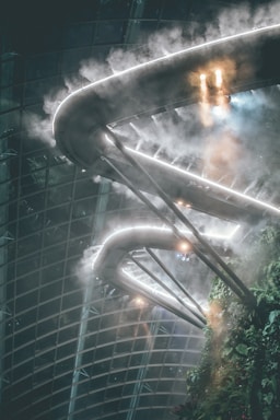 A futuristic walkway with illuminated pathways enveloped in mist, surrounded by lush greenery and structural glass panels.