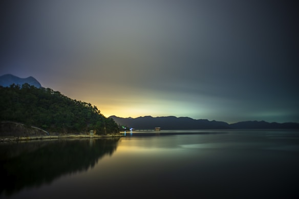 A serene lakeside landscape with a smooth, reflective body of water. In the background, there is a dense tree line and a range of silhouettes of distant mountains under an expansive sky with a gentle gradient from dark to light. A small structure is visible near the shore, emitting a faint blue light.