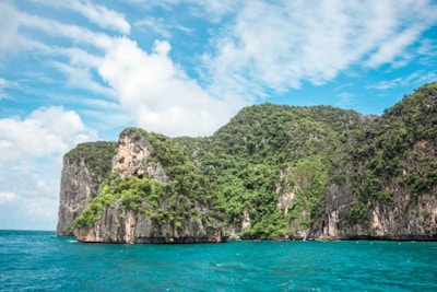 A picturesque rocky island covered with lush green vegetation rises from the turquoise blue ocean under a partly cloudy sky. The cliffs are rugged and steep, creating a dramatic and natural coastline.