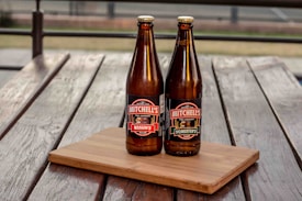 Two brown glass bottles of beer with labels that read 'Mitchell's Bosun's Bitter' and 'Mitchell's Forester's Lager' are placed on a wooden cutting board on top of a weathered wooden table. The background appears to be an outdoor setting with metal railings and a blurred outdoor space.